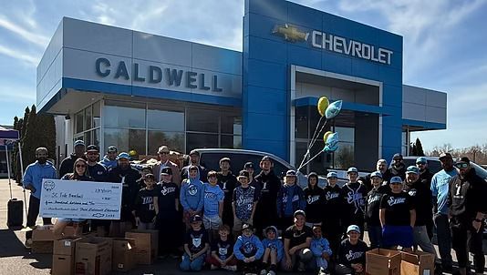 People gather at a Chevrolet dealership. A group holds a large check, with balloons.