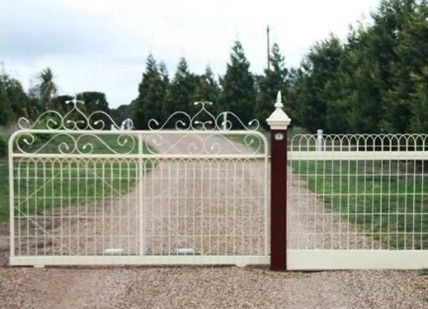 A white gate stands besides a wooden fence, leading to a dirt road surrounded by greenery.