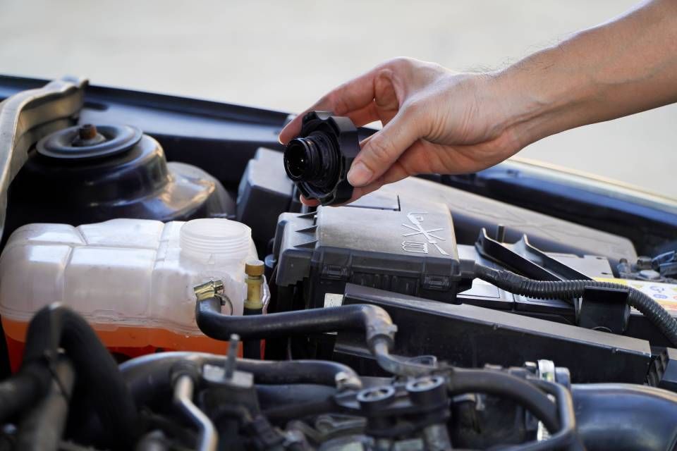 Hand removing black cap from a car engine, next to coolant reservoir.