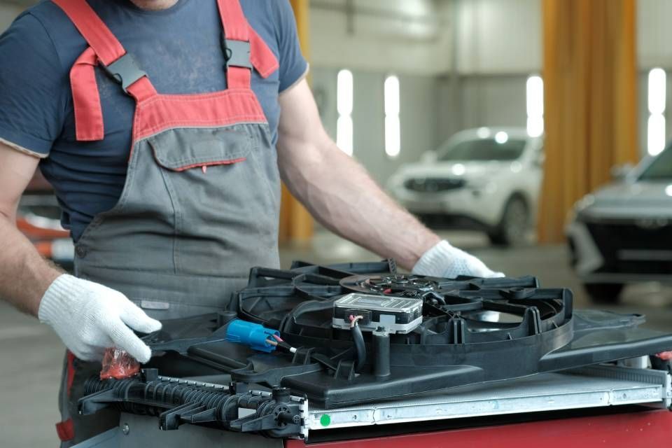 Mechanic in overalls and gloves holding car cooling fan in a repair shop.
