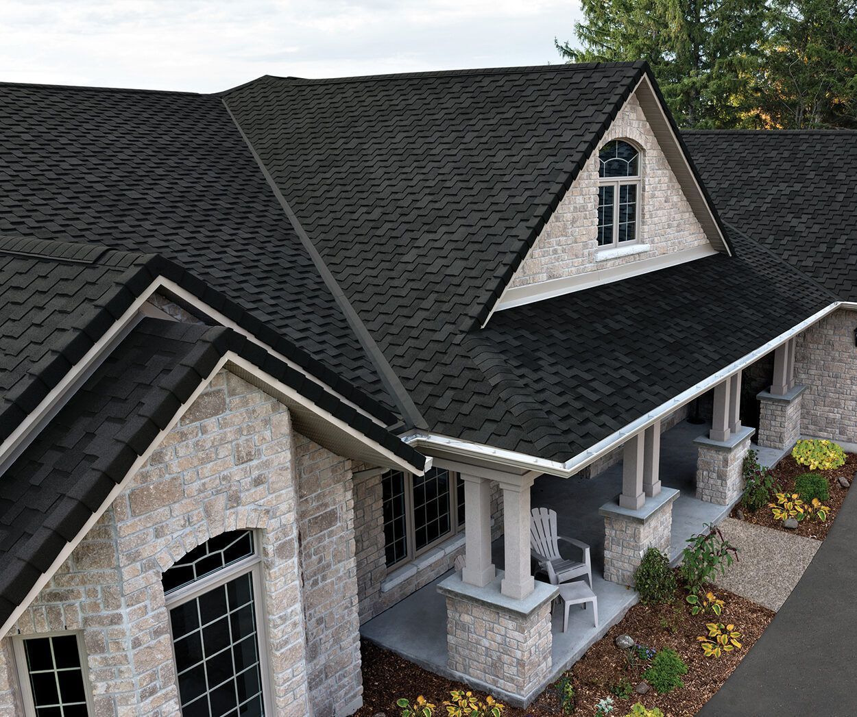 Aerial view of a stone house with dark shingle roof, dormers, and a covered front porch