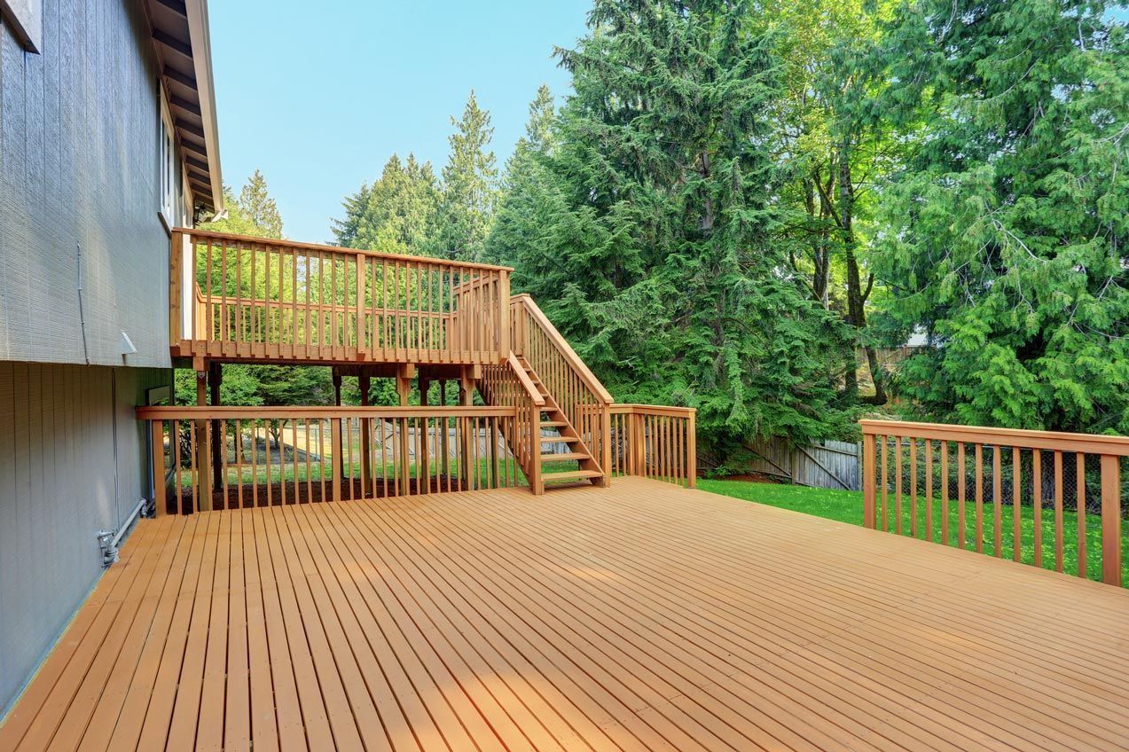 Large wooden deck with stairs and railings beside a house, surrounded by trees.
