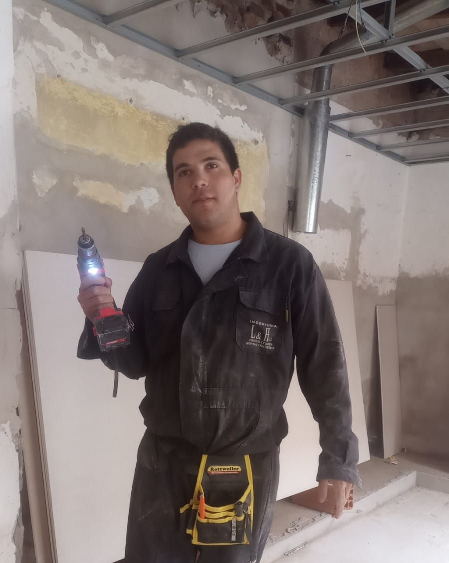 Un obrero de la construcción sostiene una botella en una habitación sin terminar con vigas de techo a la vista y paredes remendadas.