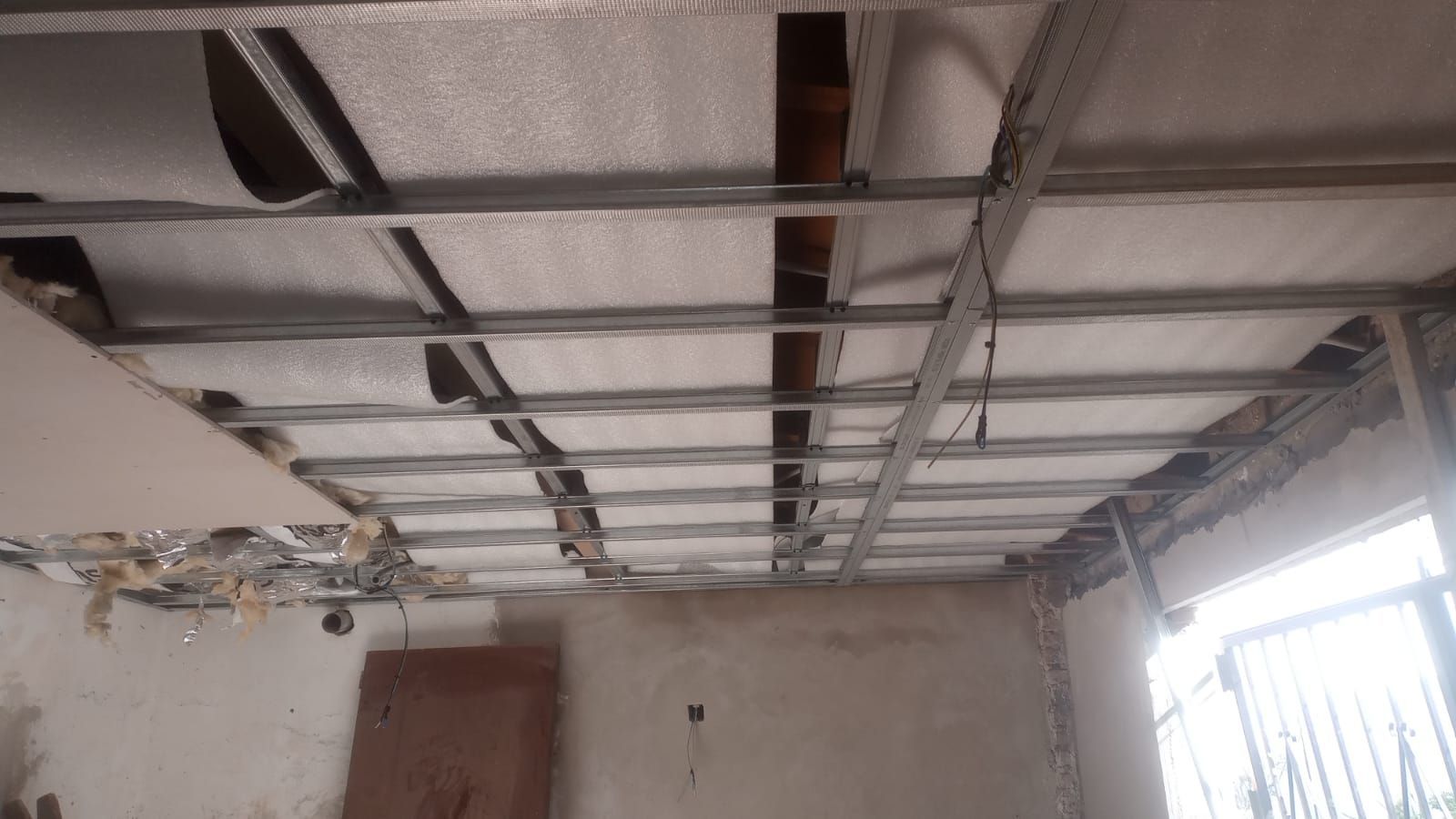 Techo parcialmente construido con estructura metálica, aislamiento a la vista y una ventana luminosa en una habitación vacía.