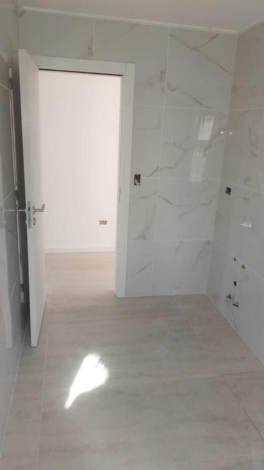 Habitación vacía con azulejos, paredes blancas, puerta abierta y luz solar que ilumina el suelo de madera clara.
