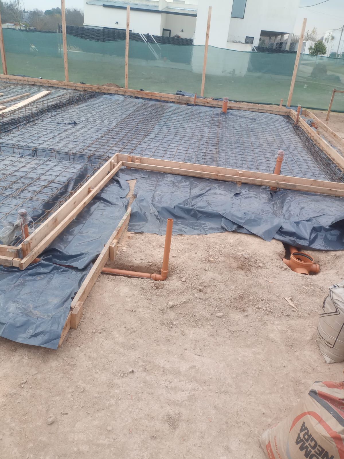 Obra en construcción con estructura de cimientos cubierta de plástico negro y suelo de grava detrás de una valla de seguridad.
