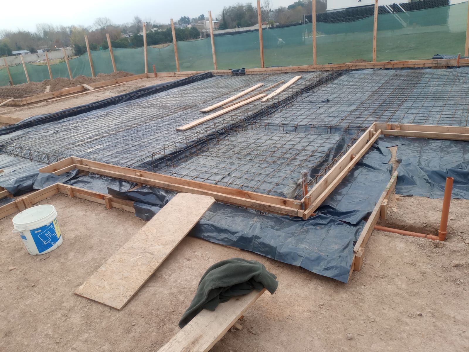 Obra en construcción con malla de barras de refuerzo y encofrado de madera extendido sobre lona negra en terreno arenoso.