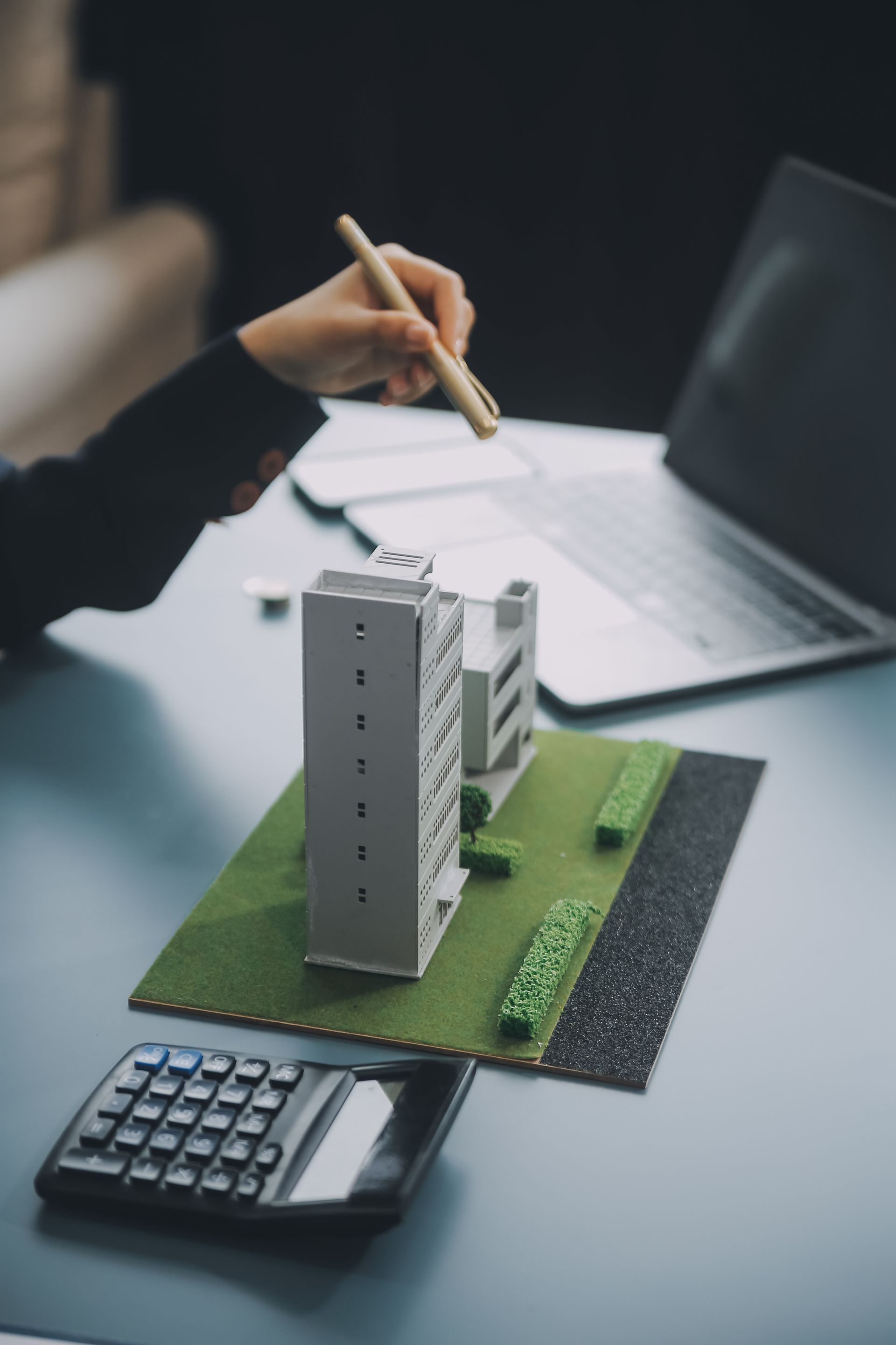 Hand holding a pencil above a scale model of a building on a desk with a calculator and laptop nearby
