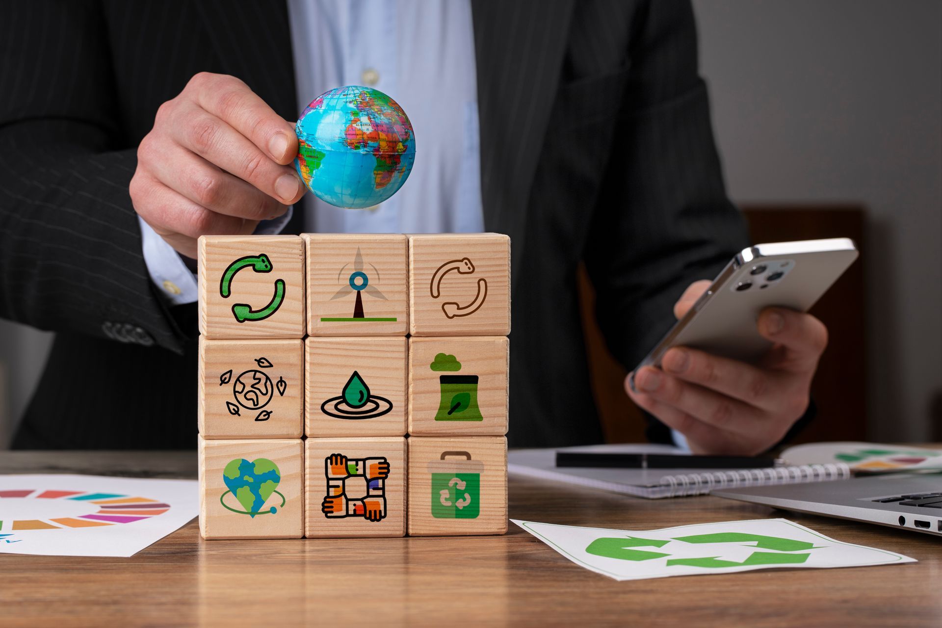 Person holding globe above blocks with sustainability icons, using phone.