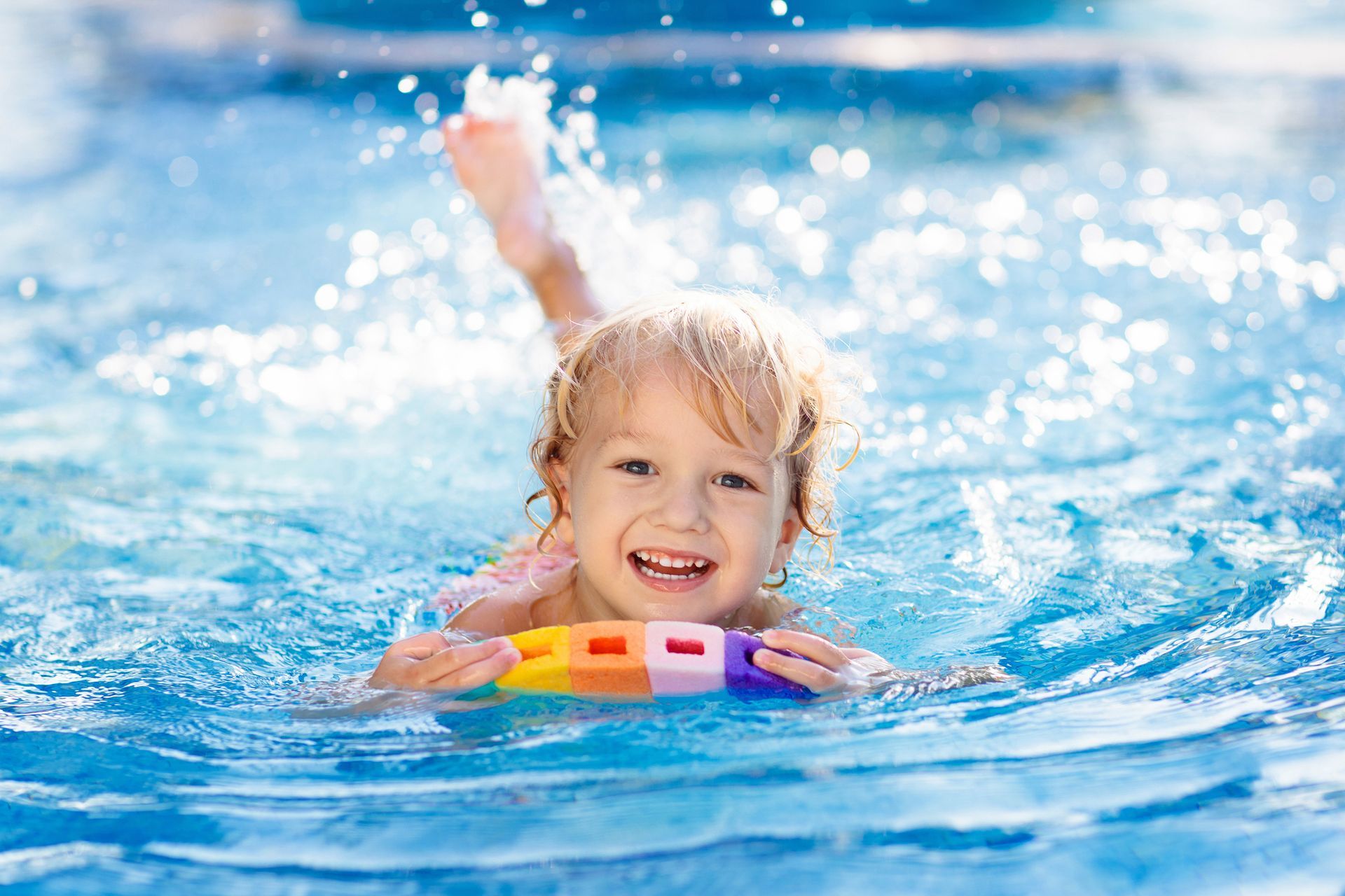 Child swimming in a pool, holding a colorful float, smiling, with water splashing around.