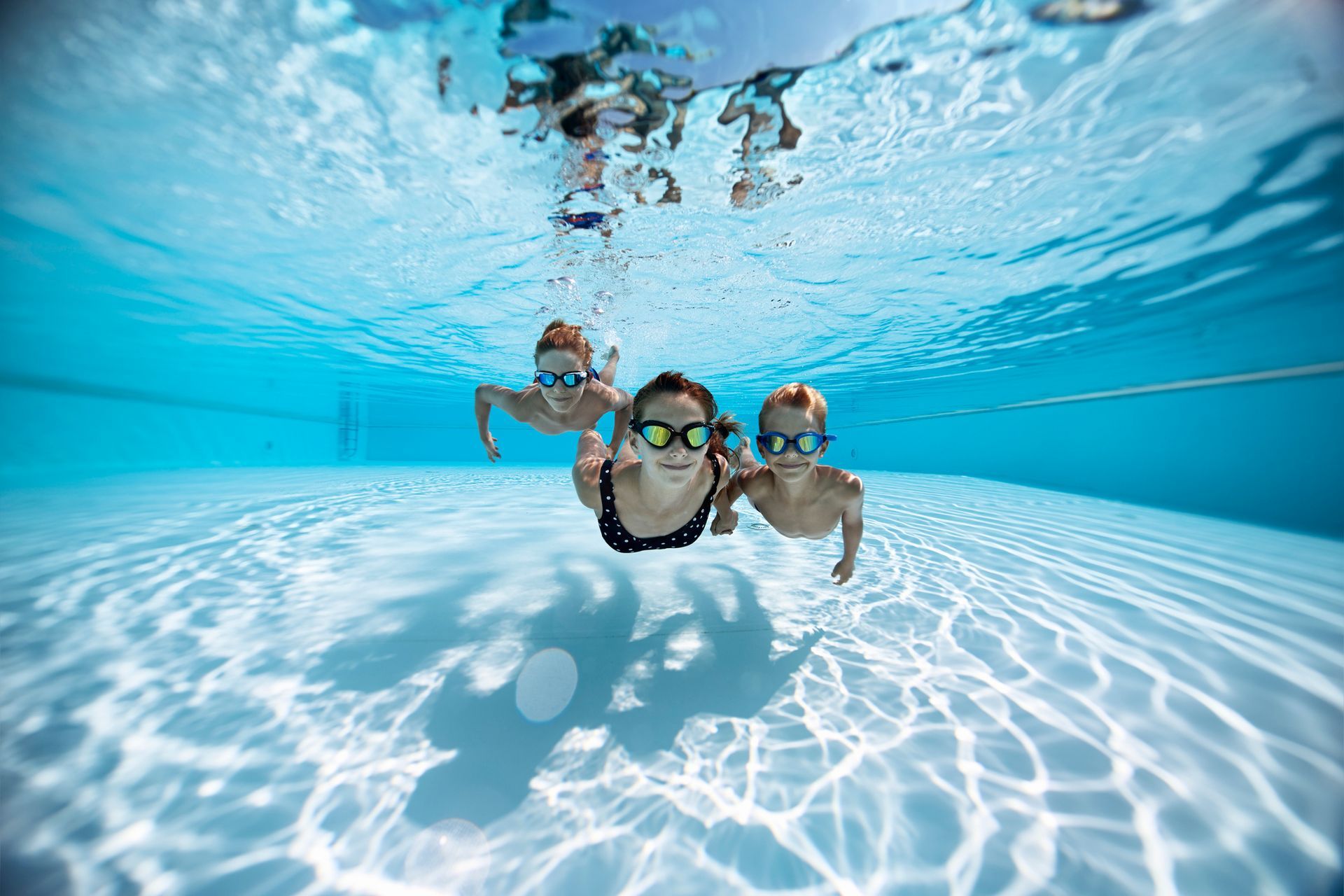 Three children swimming underwater in a pool, wearing goggles.