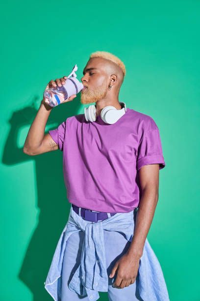 Man drinking from water bottle, wearing purple shirt and light blue jacket, headphones around his neck, green background.