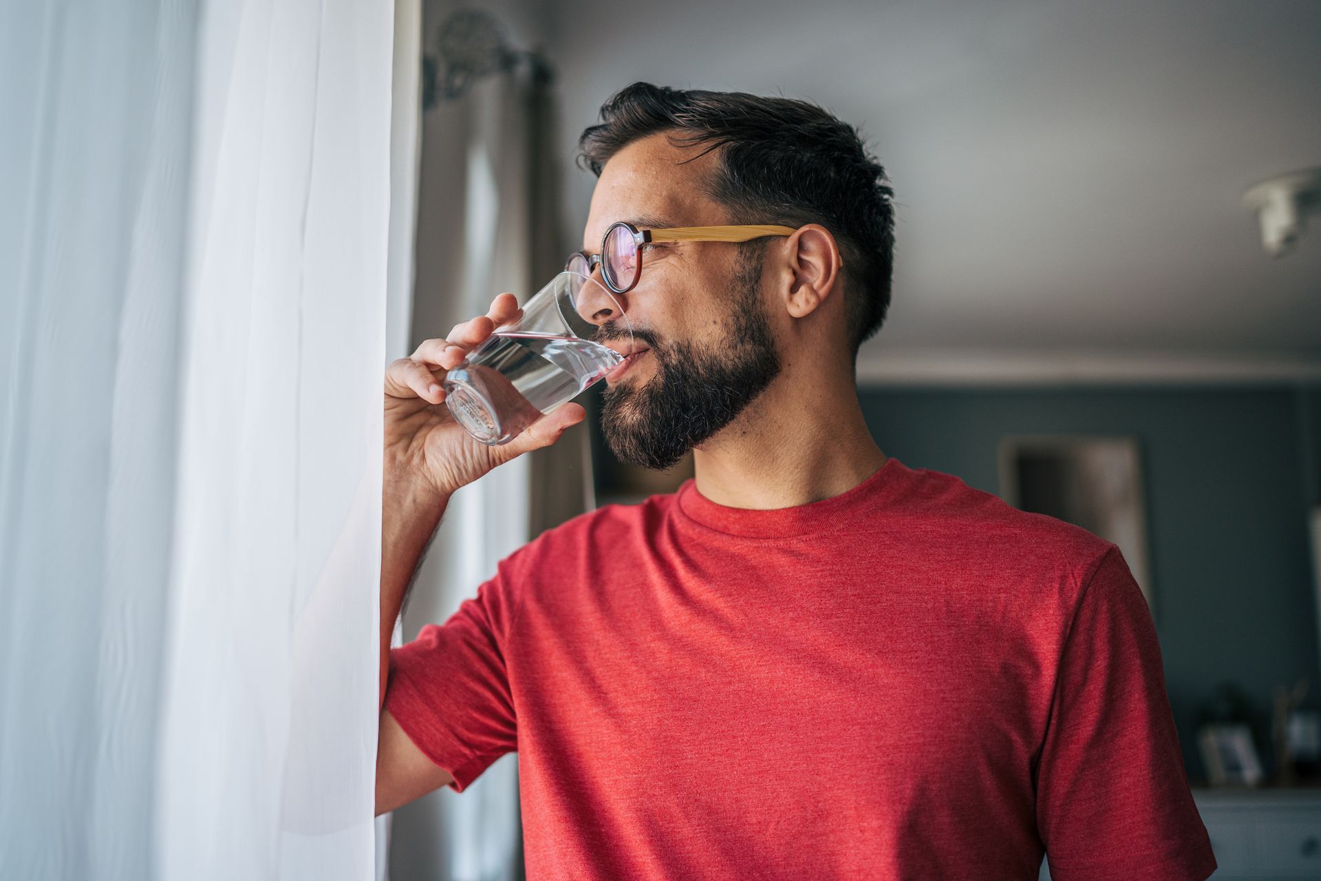 Man in red shirt drinks water from a glass, looking out a window.