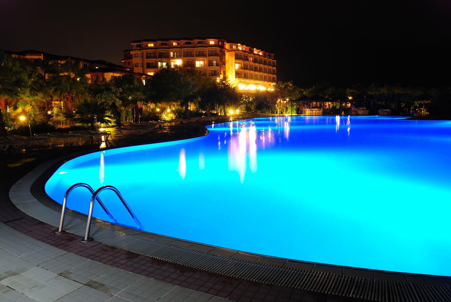 Night view of a large, glowing blue pool with a building in the background. Pool ladder visible.