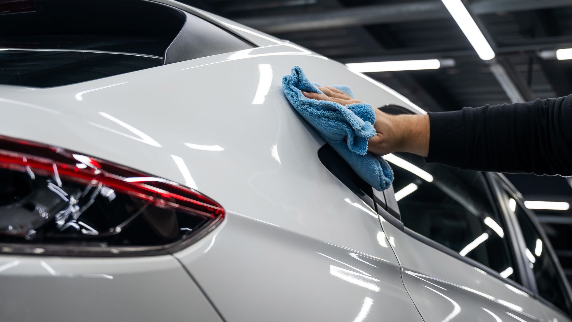 A person cleans a white car with a blue microfiber cloth inside a brightly lit auto detailing shop.