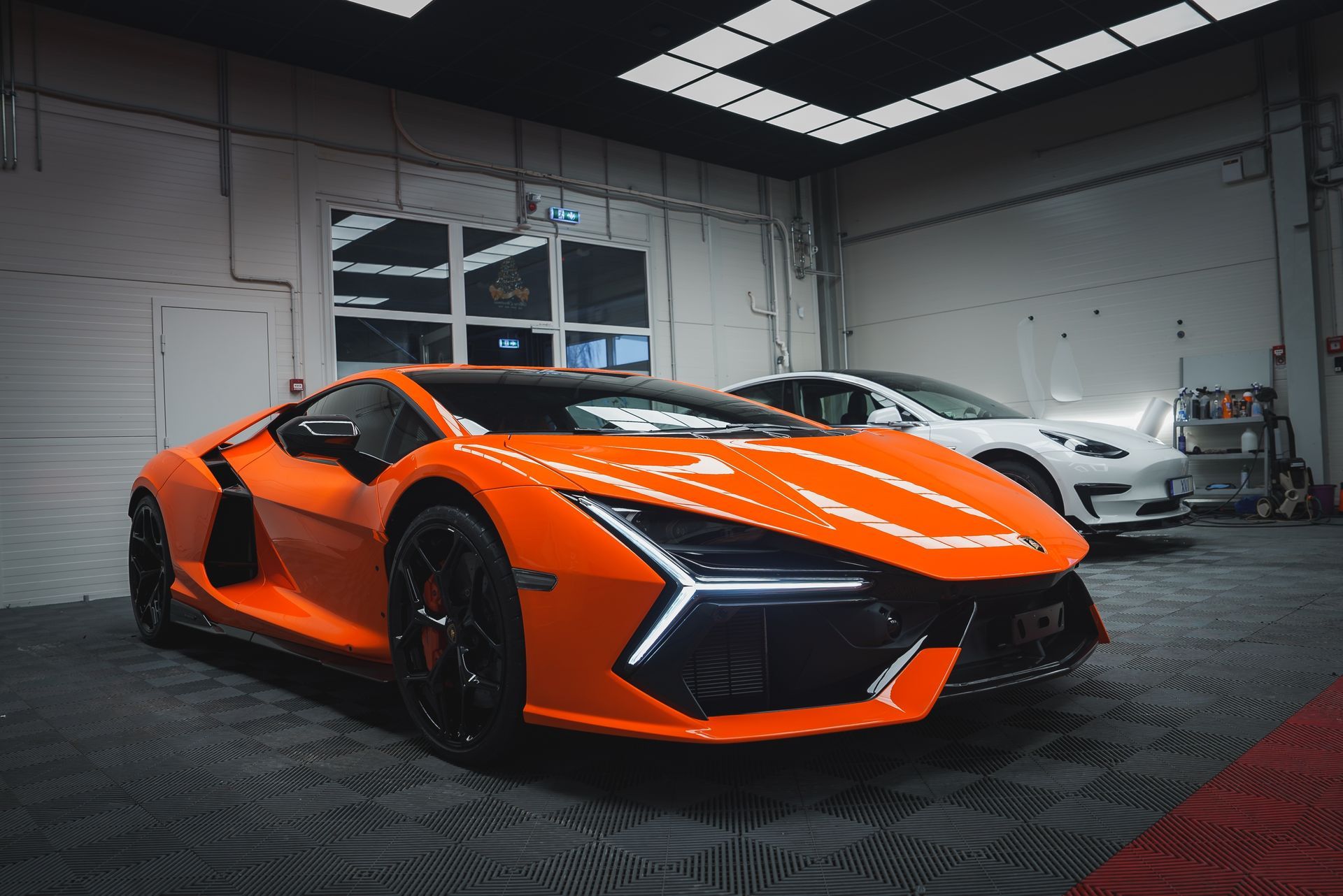 An orange Lamborghini Revuelto parked in a brightly lit indoor garage, with a white car visible in the background.