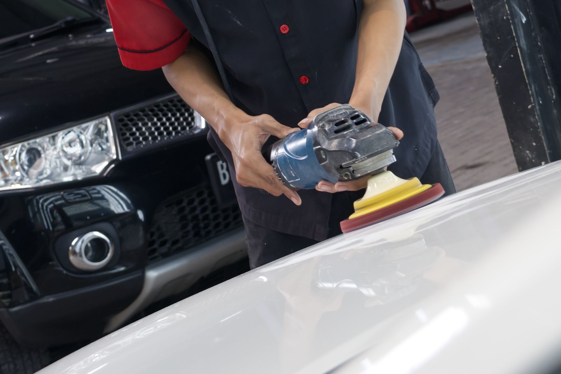 A worker uses a power buffer to polish the white hood of a car in a garage next to a black vehicle.