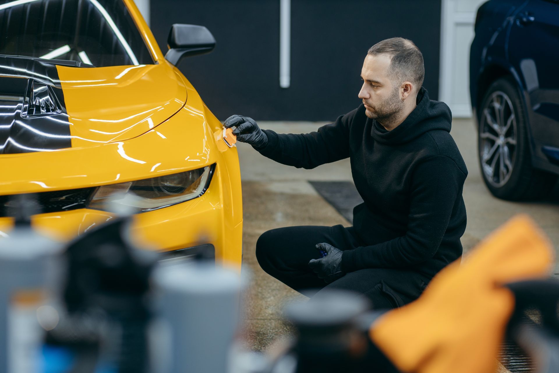 A person in black clothing wearing gloves inspects the paint on a yellow car in a professional garage setting.