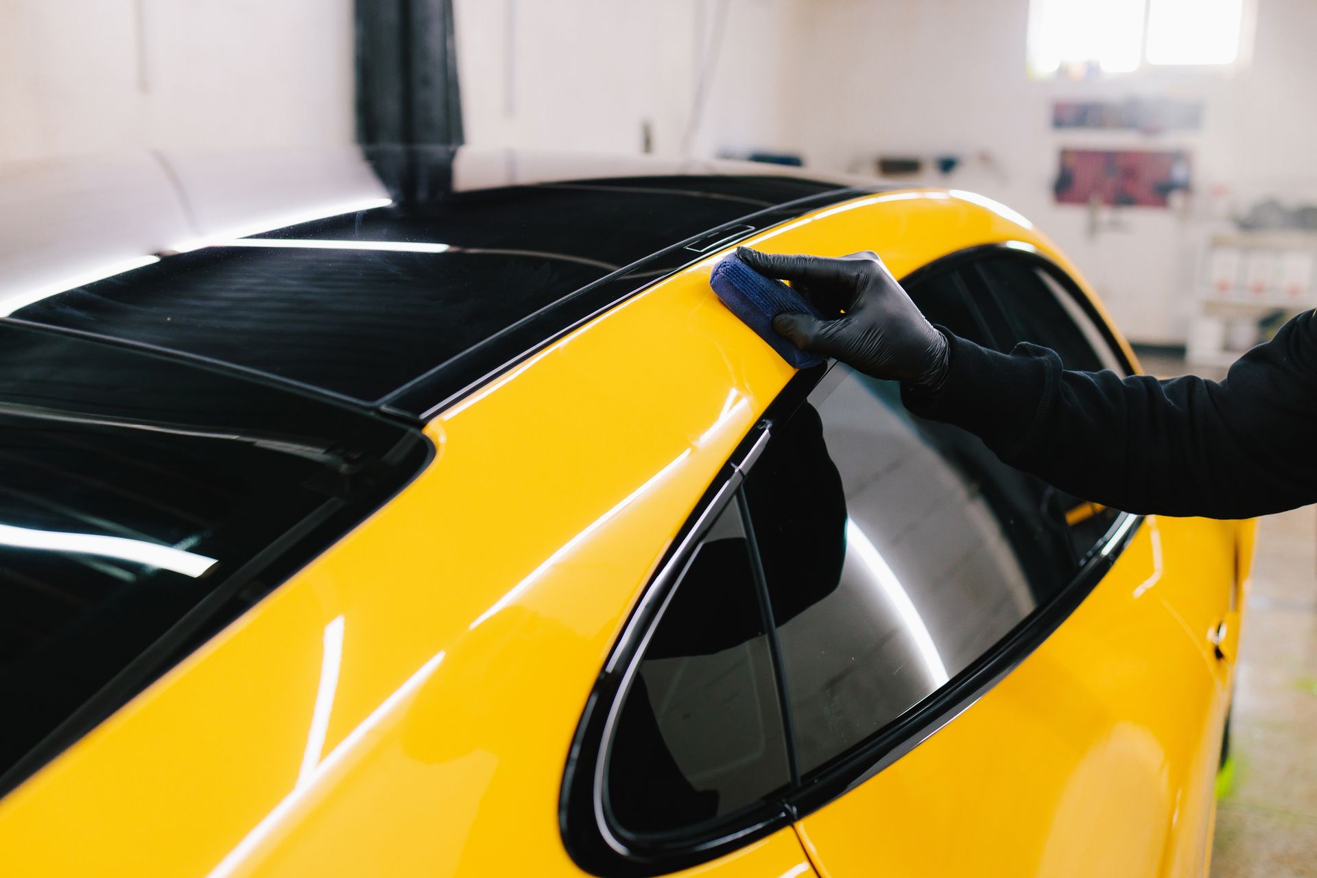 A gloved hand uses a blue applicator pad to detail the yellow exterior of a car in a shop.