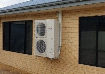 Air conditioner mounted on a tan brick wall between two windows.