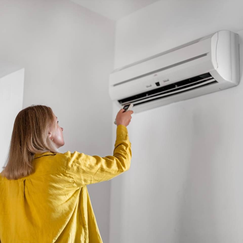 Woman in yellow shirt operating wall-mounted air conditioner with a remote in a white room.