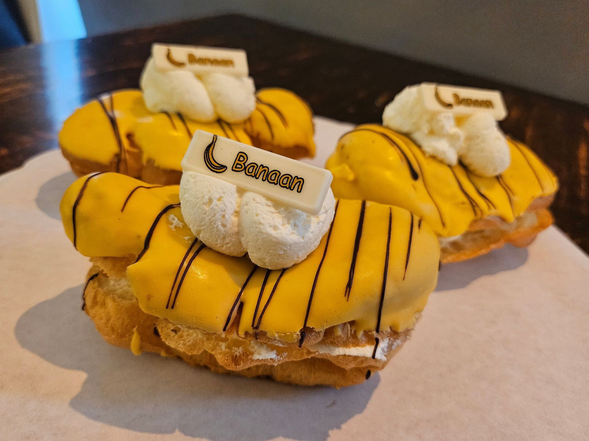 Drie banaanvormige soezen met felgele glazuur, chocoladesaus, slagroom en chocoladeblokjes met logo.