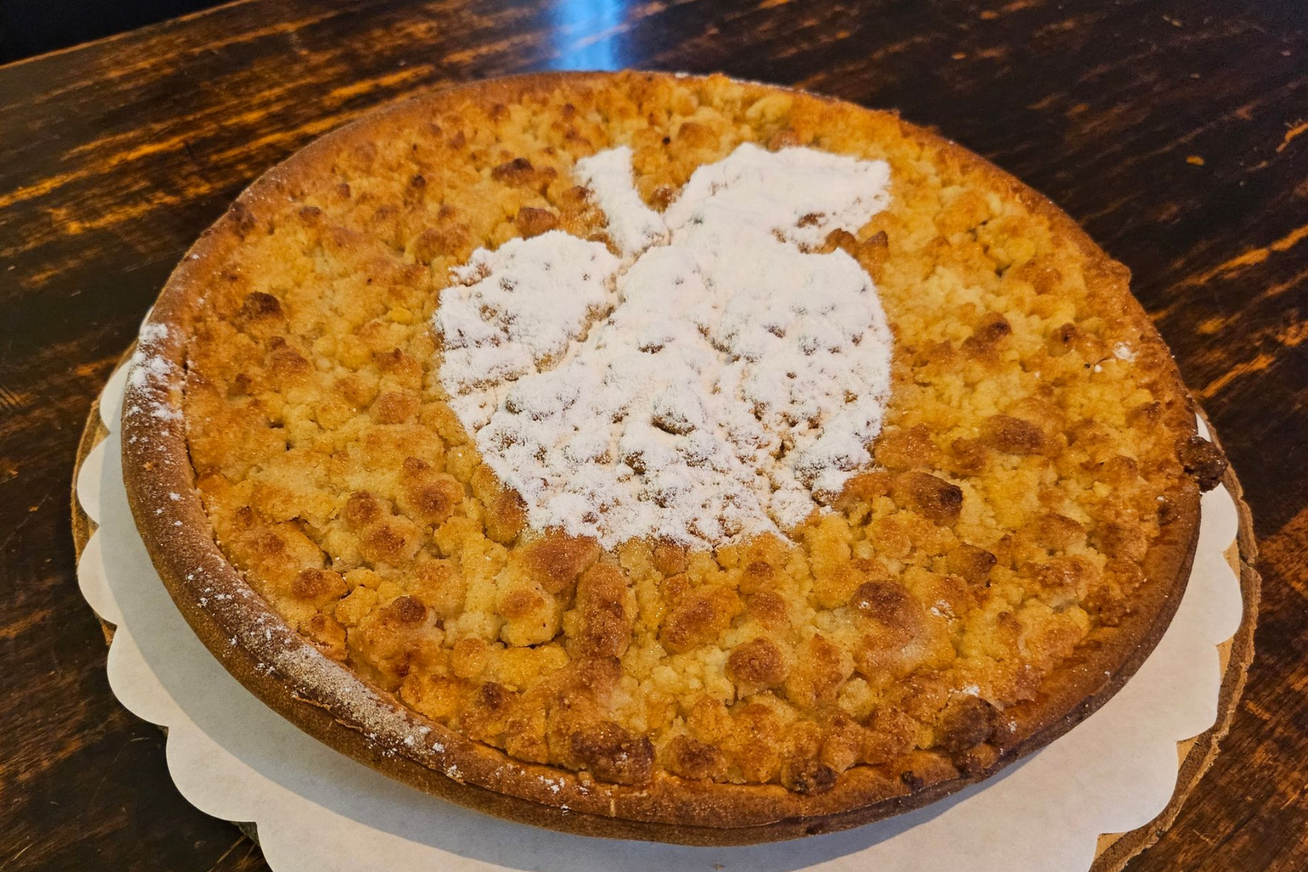 Een ronde appelcrumbletaart op een gegolfde papieren bodem, met poedersuiker in de vorm van een appel in het midden.