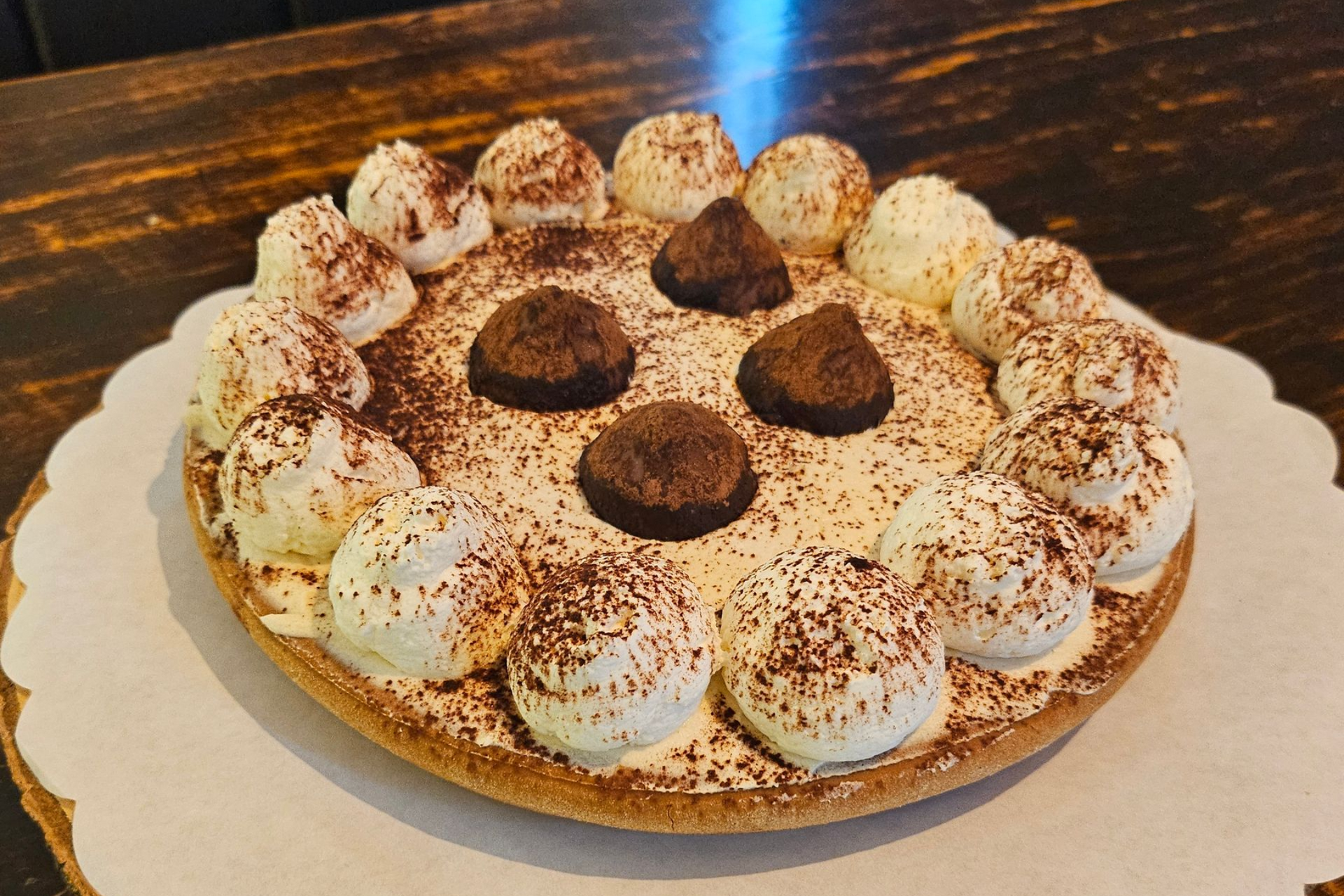 Een ronde taart, bedekt met toefjes slagroom, bestrooid met cacaopoeder en versierd met vier chocoladetruffels.