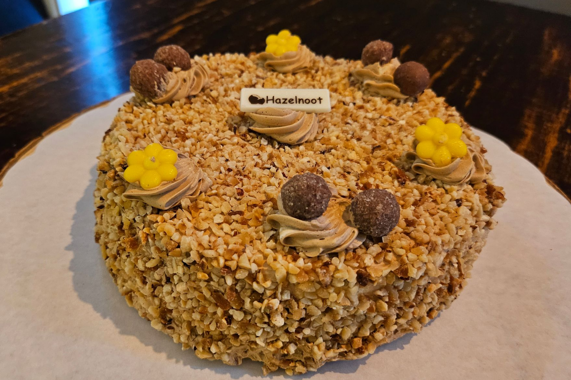 Een met hazelnoten bedekte taart, versierd met chocoladebolletjes, gele glazuurbloemen en een klein bordje.
