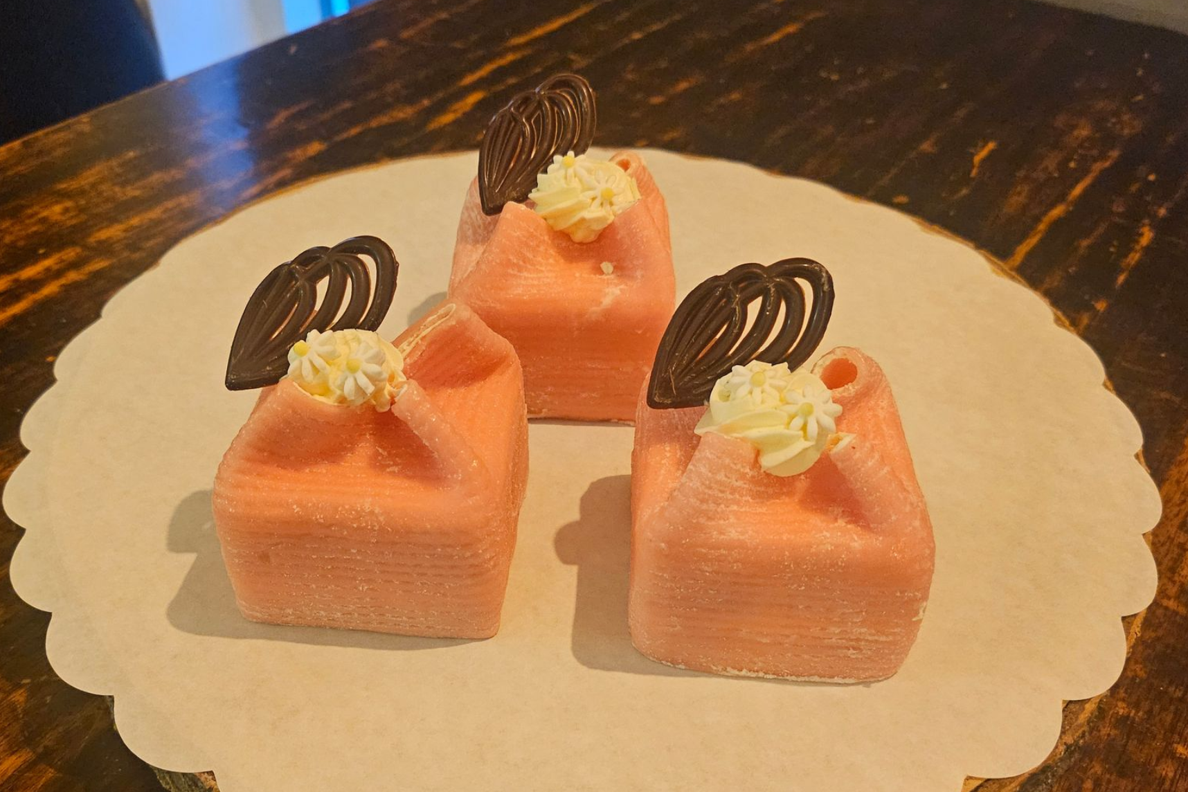 Drie kleine, vierkante, roze cakejes met glazuur, afgewerkt met slagroom en versierd met pure chocolade.