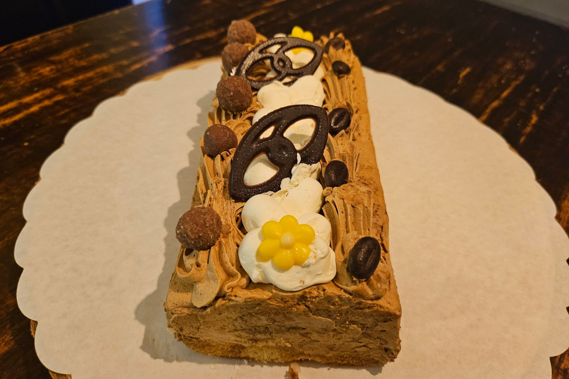 Een rechthoekig dessertrolletje met koffiesmaak, versierd met slagroom, stukjes chocolade en een bloem.