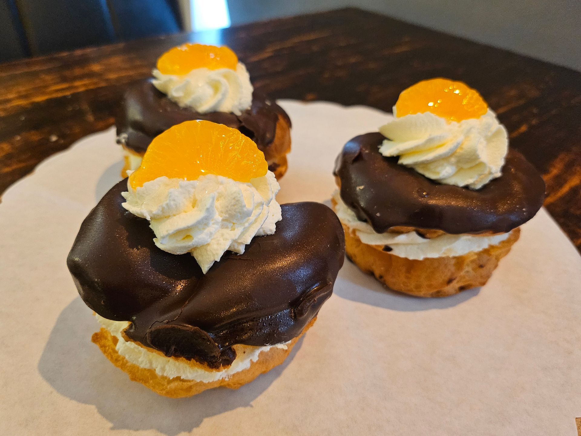 Drie met crème gevulde gebakjes, gegarneerd met pure chocoladeglazuur, slagroom en een schijfje mandarijn.