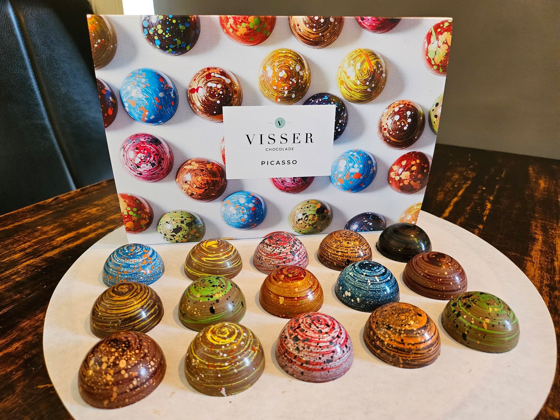 Een assortiment kleurrijke chocoladebonbons met verschillende patronen, daarachter een doos met het merklogo.