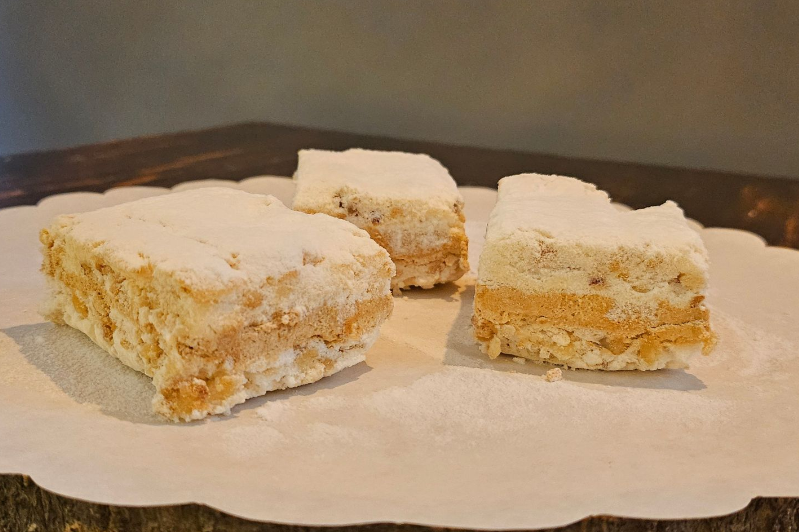 Drie vierkante stukken gelaagd dessert met een zandkleurige kern en witte, poederachtige toppings.