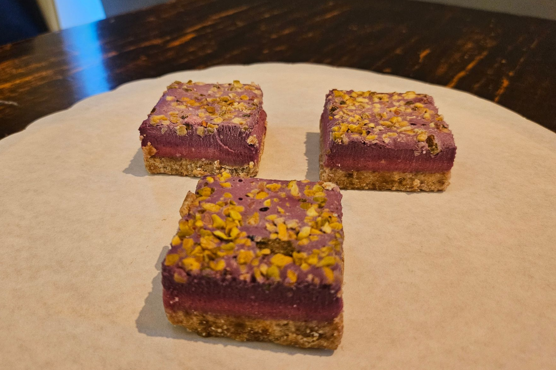 Drie vierkante gebak met een nootachtige bodem, een donkere bessenkleurige vulling en een topping van gemalen pistachenoten.