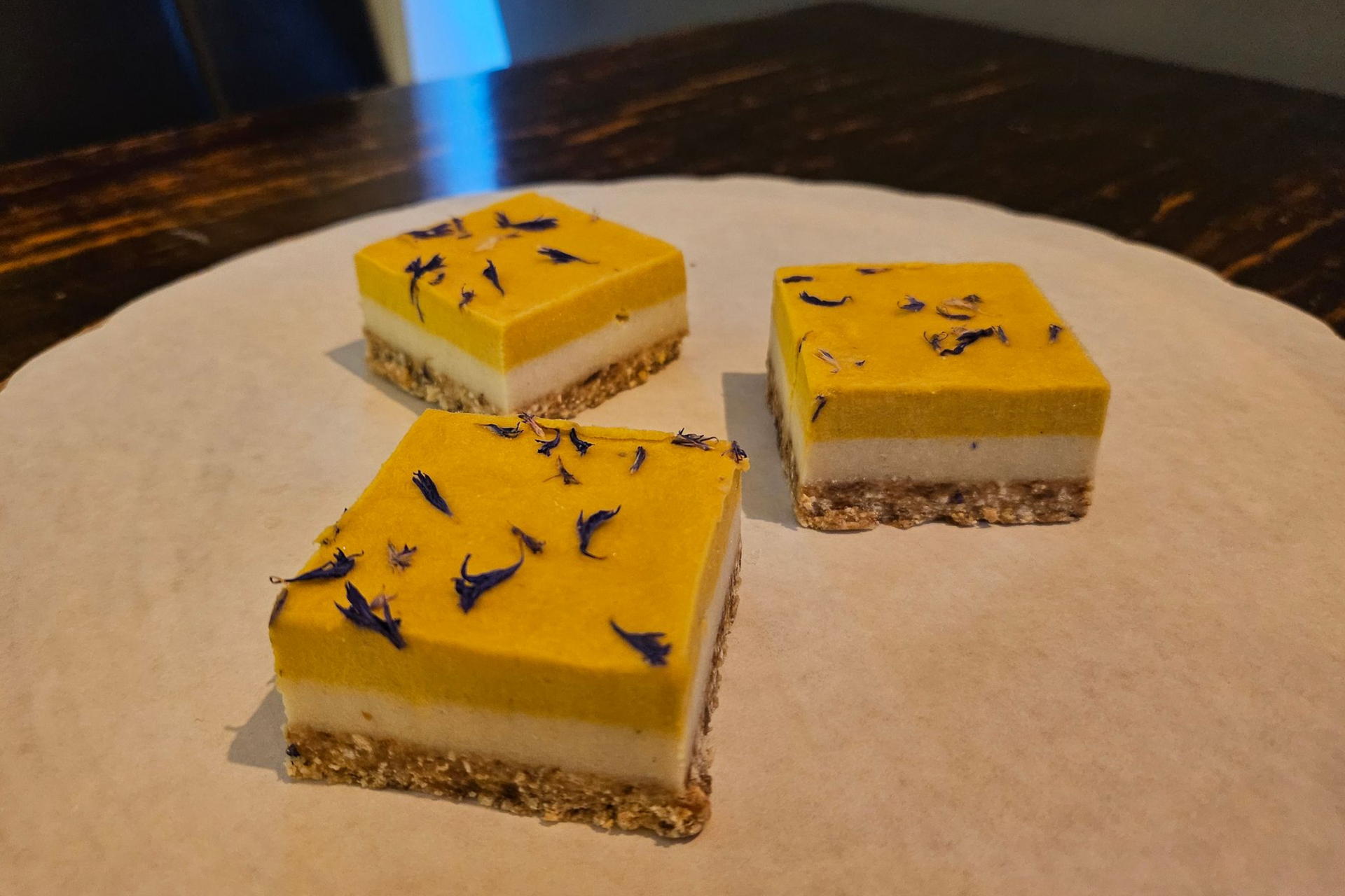 Drie vierkante, gelaagde dessertrepen met een notenkorst, een witte crèmevulling en een felgele topping.