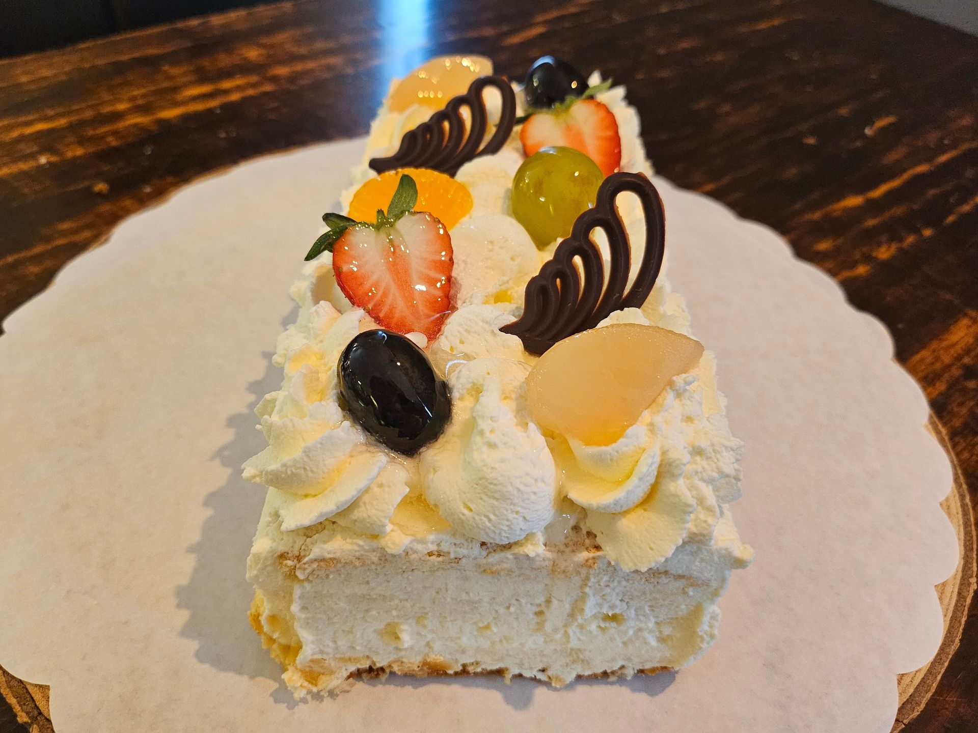 Een rechthoekige slagroomtaart, versierd met aardbeien, druiven, perzikschijfjes en decoratieve chocoladekrullen.