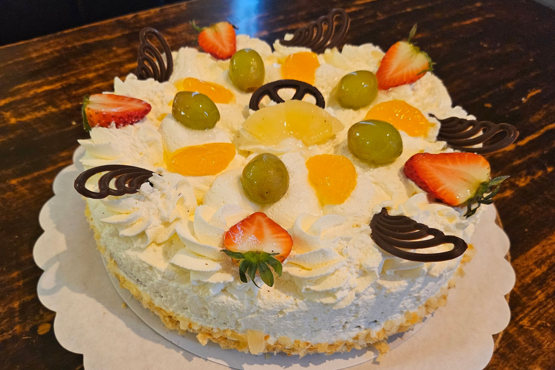 Een ronde vruchtentaart, gegarneerd met slagroom, aardbeien, druiven, perziksnippers en decoratieve chocoladekrullen.