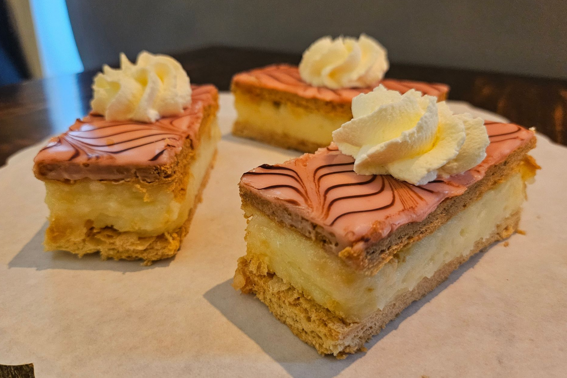 Drie rechthoekige plakken mille-feuille gebak met roze geglazuurde bovenkant, toefjes slagroom en gelaagde vulling.
