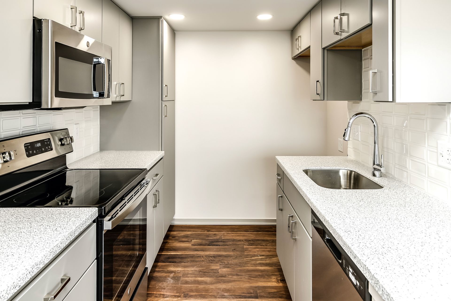 Bright narrow kitchen with white cabinets, stainless appliances, dark wood floor, and a sink counter on the right.