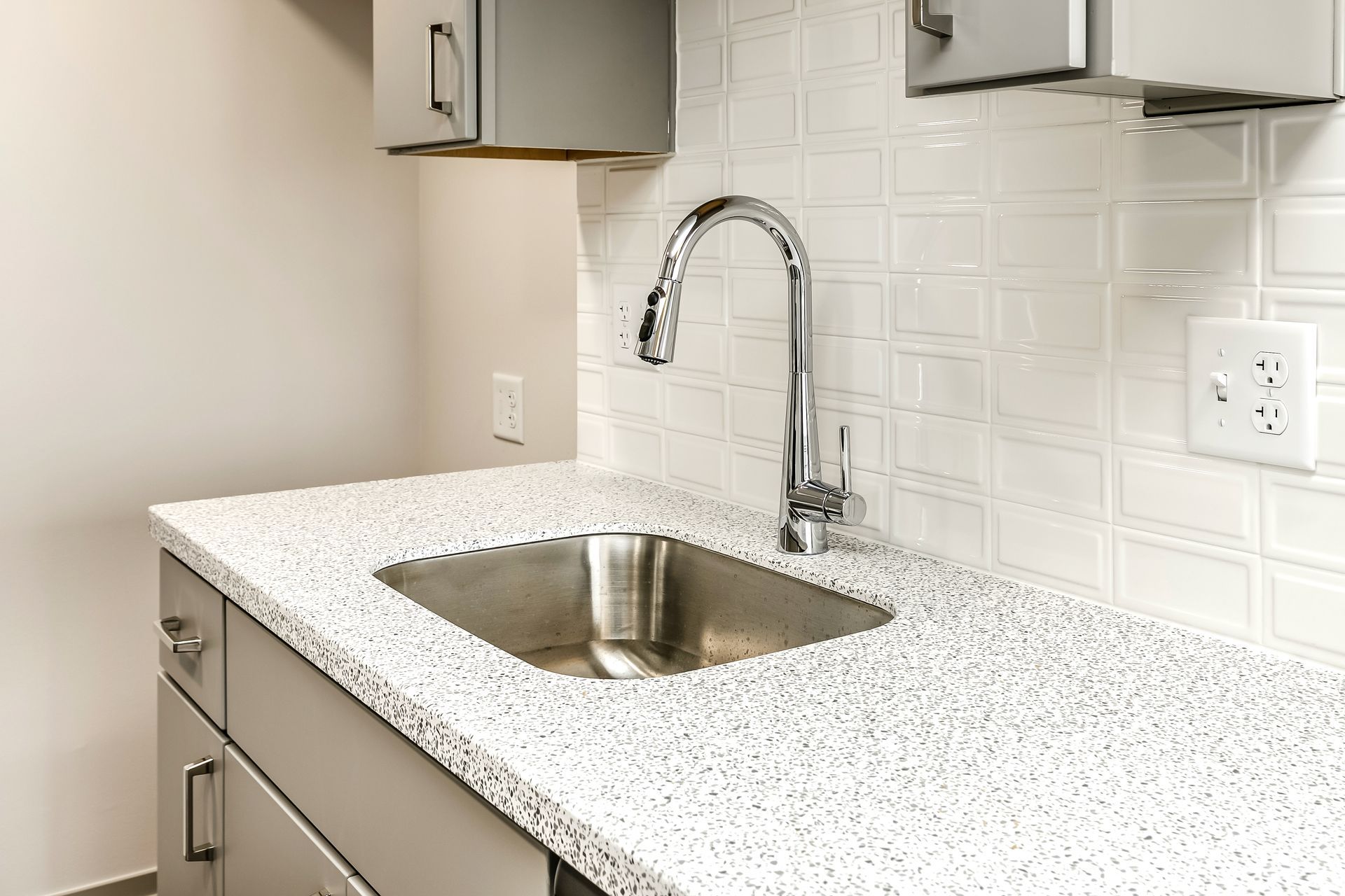 Modern kitchen sink with chrome faucet, speckled countertop, and white tile backsplash.