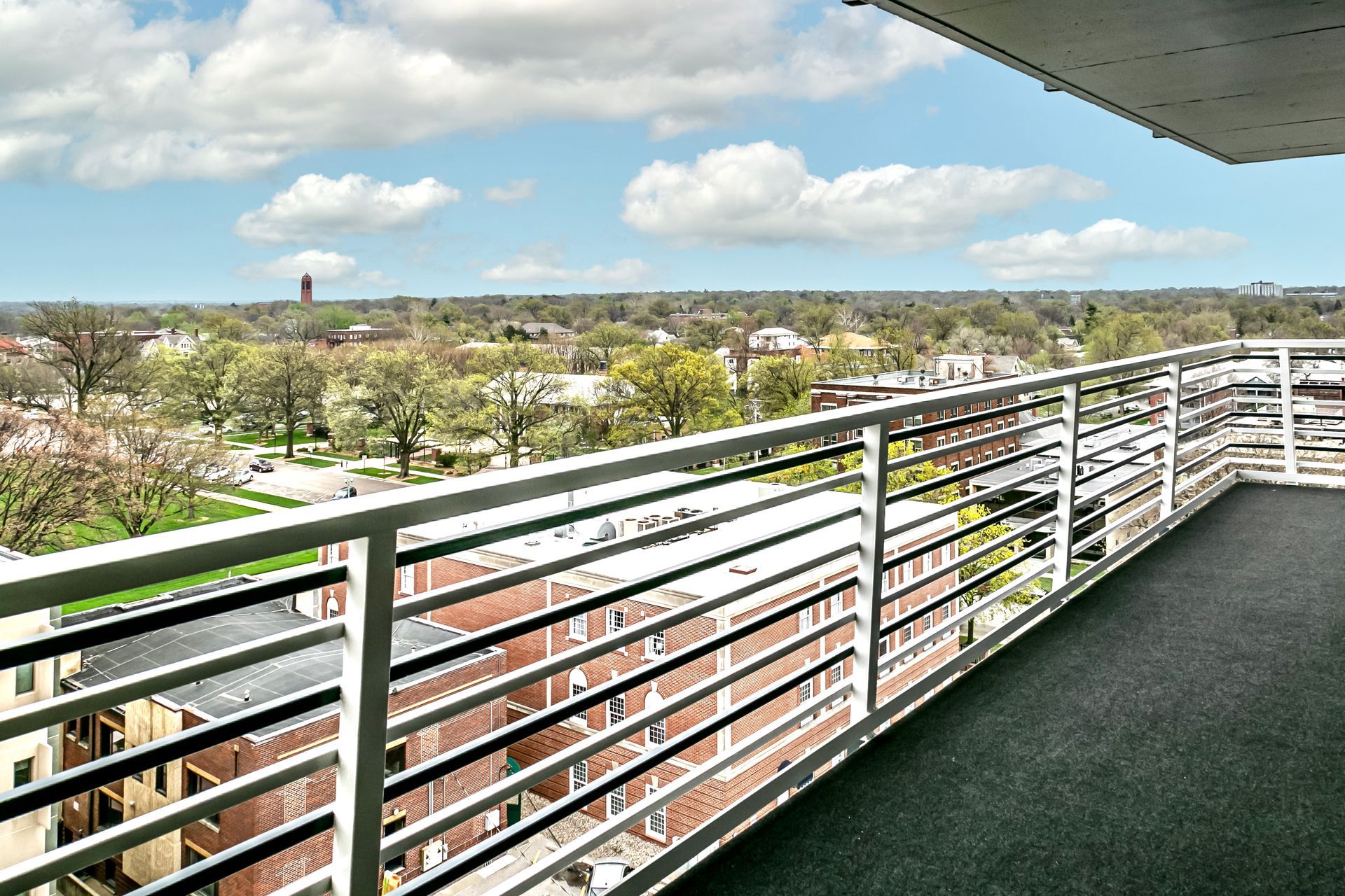 Balcony view over tree-lined neighborhood with white railing, cloudy sky, and distant tower