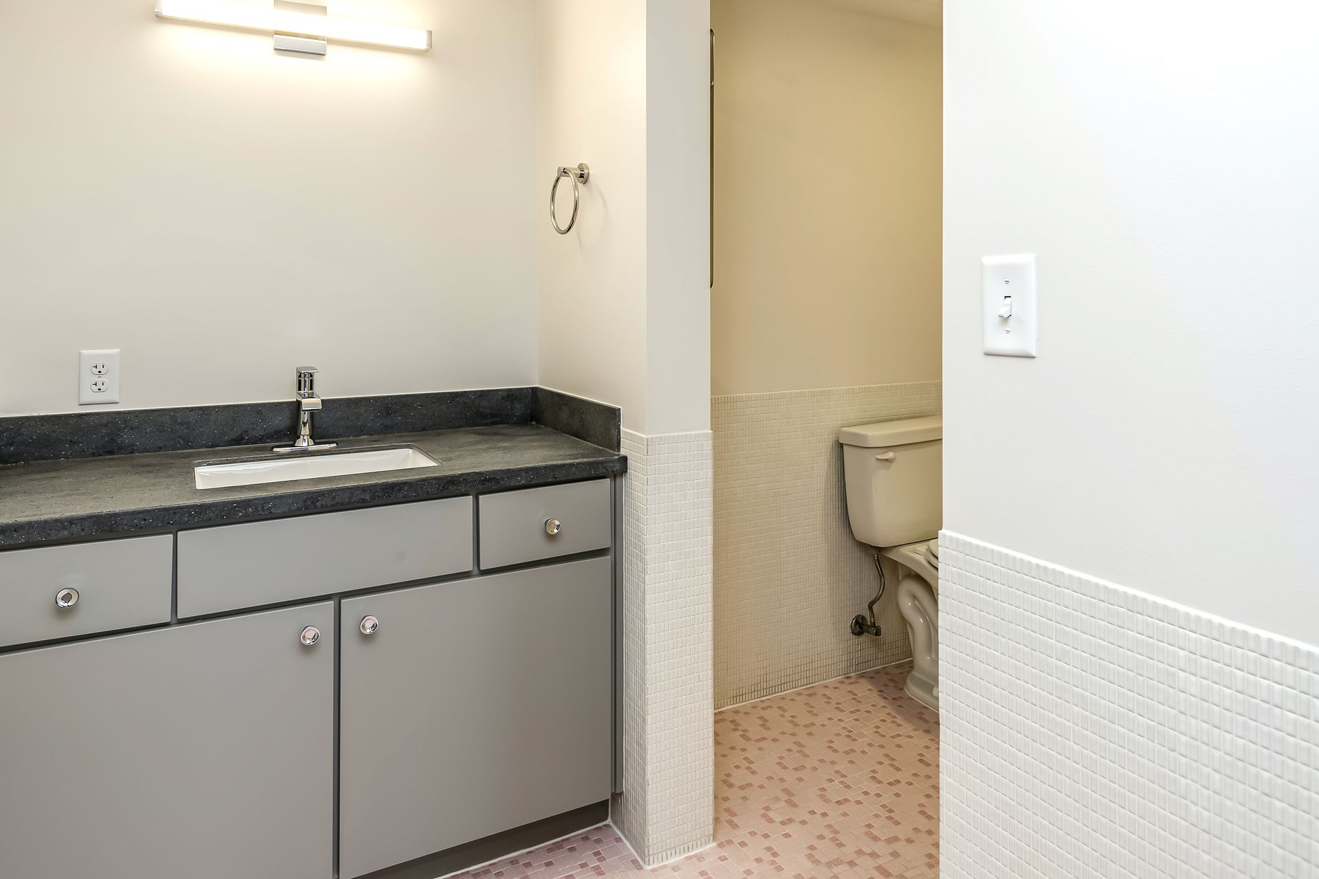 Small bathroom vanity with gray cabinets, black countertop, sink, and toilet visible in the next stall.