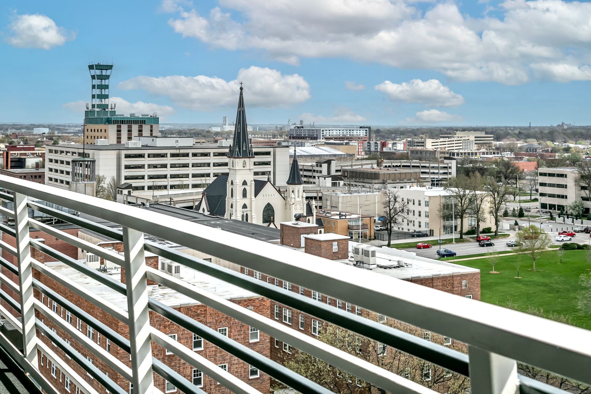 City skyline viewed from a balcony, with a church steeple, apartment buildings, and a green field under a cloudy blue sky