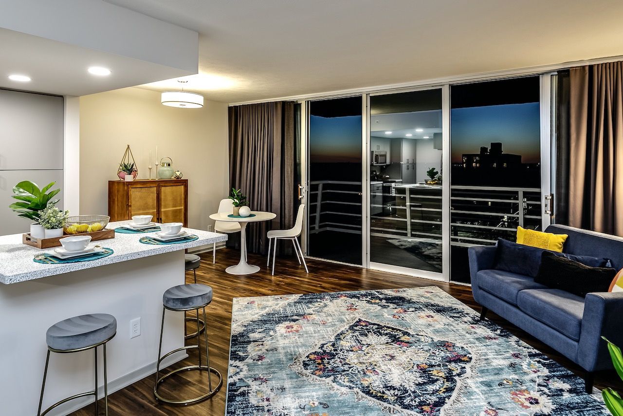Modern living room with balcony view, blue sofa, round rug, kitchen island, and warm lighting
