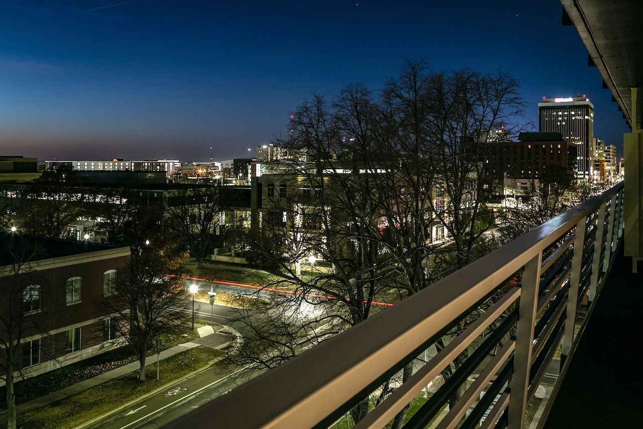 City skyline at dusk viewed from a balcony, with lit buildings, trees, and a blue evening sky