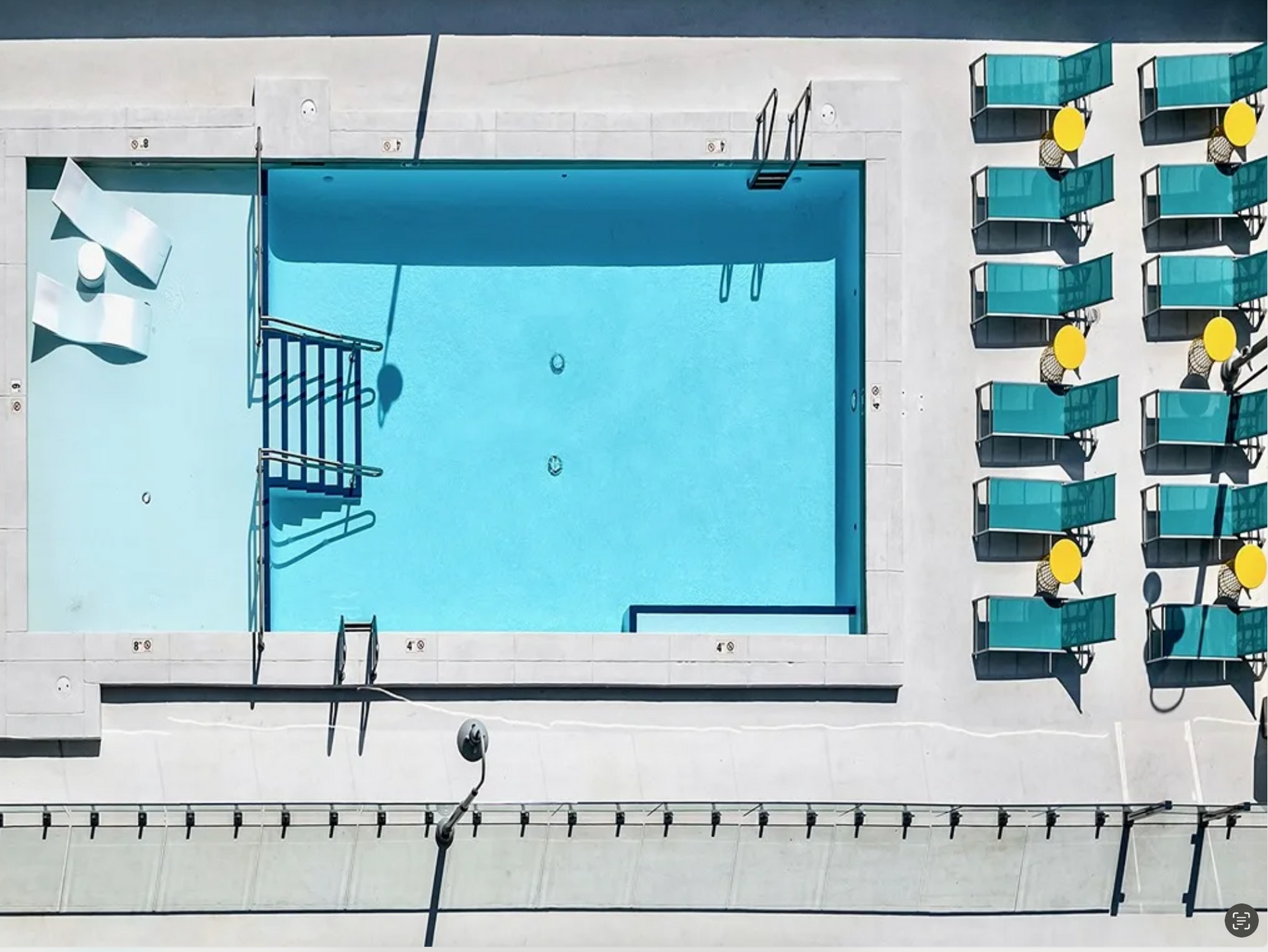 Overhead view of a blue swimming pool with lounge chairs and tables on a white deck.
