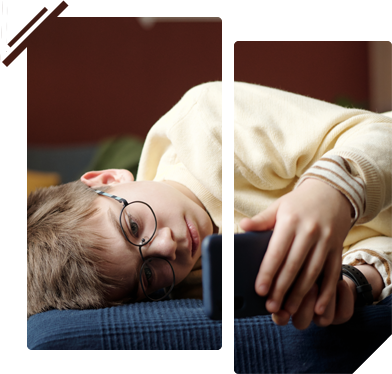 A young boy wearing glasses is laying on a bed looking at a cell phone