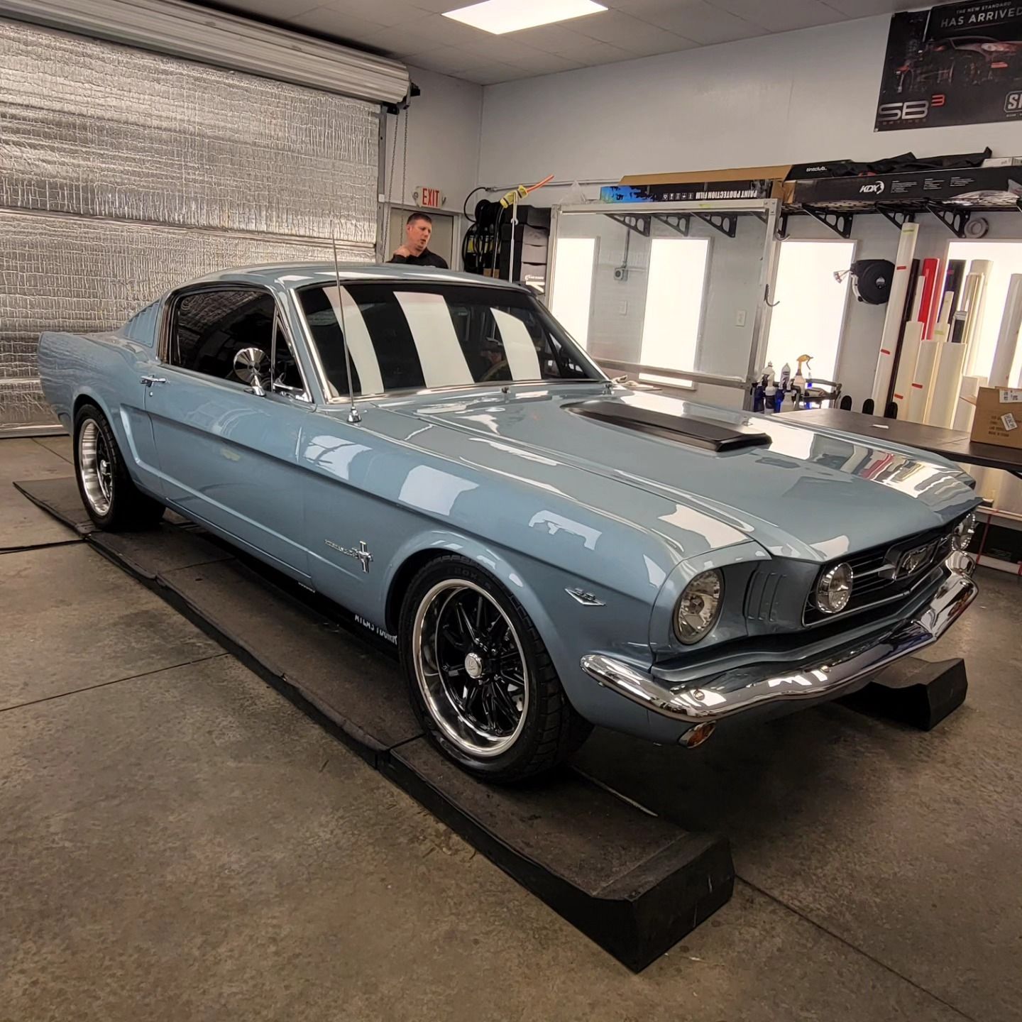 A light blue classic Ford Mustang fastback parked on a ramp in a garage with a person visible in the background.