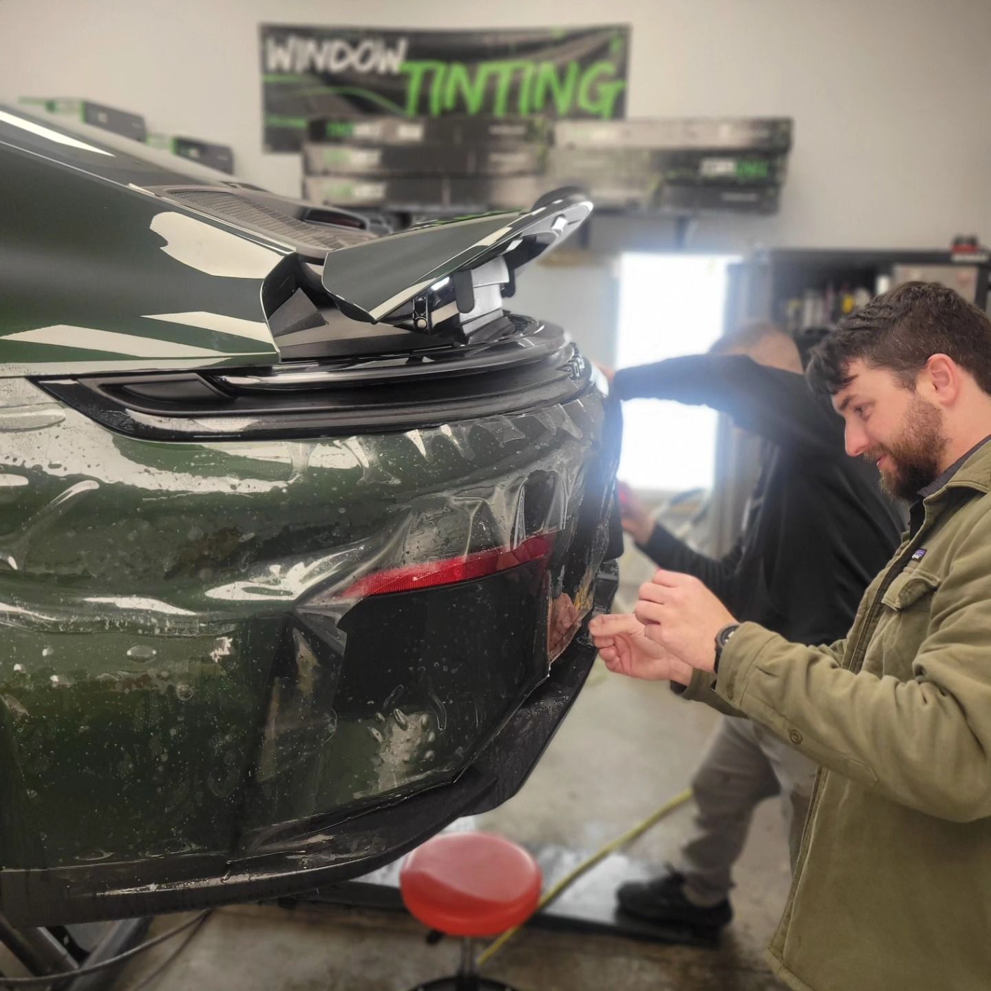 Hands applying tinted film to a car's rear window in a garage.
